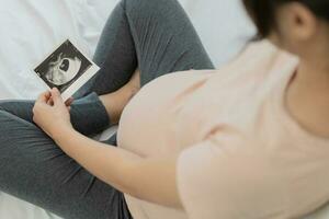 zwanger vrouw is op zoek Bij een echografie foto van foetus. moeder voorzichtig accenten de baby Aan maag. Vrolijk, familie, groei, zwangerschap, genot , bereiden pasgeboren, nemen zorg, gezondheidszorg, buik.
