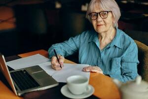 mooi volwassen senior vrouw met bril zit Bij een tafel in voorkant van een laptop ongewijzigd foto