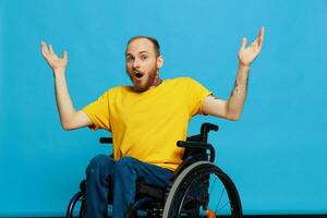 een Mens zit in een rolstoel bedachtzaamheid in een t-shirt Aan een blauw achtergrond in de studio, verheven zijn handen omhoog en geopend zijn mond in verrassing, getatoeëerd lichaam foto