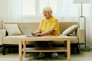 gelukkig ouderen vrouw met laptop typen in hoofdtelefoons glimlach zittend Bij huis video telefoongesprek, helder modern interieur, levensstijl online communicatie. foto