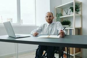 een Mens in een rolstoel, een zakenman in de kantoor werken Bij een laptop, schrijven naar beneden een plan in een notitieboekje, bedachtzaamheid, integratie in samenleving, de concept van werken een persoon met handicaps foto