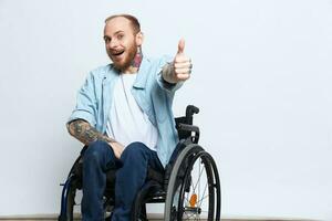 een Mens in een rolstoel looks Bij de camera shows een duimen omhoog, geluk, met tatoeages Aan zijn handen zit Aan een grijs studio achtergrond, Gezondheid concept Mens met handicaps foto