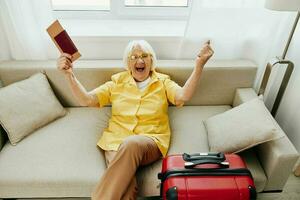 gelukkig senior vrouw met paspoort en reizen ticket Ingepakt een rood koffer, vakantie en Gezondheid zorg. glimlachen oud vrouw blij zittend Aan de sofa voordat de reis verheven haar handen omhoog in vreugde. foto