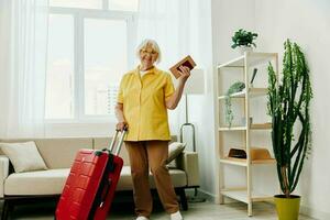 gelukkig senior vrouw met paspoort en reizen ticket Ingepakt een rood koffer, vakantie en Gezondheid zorg. glimlachen oud vrouw blij staat in de huis voordat de reis. foto