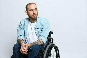 een Mens in een rolstoel looks bedachtzaam Bij de camera, kopiëren ruimte, met tatoeages Aan zijn armen zit Aan een grijs studio achtergrond, de concept van Gezondheid een persoon met handicap, een echt persoon foto