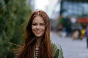 portret van een mooi jong meisje in de stad op zoek in de camera glimlach met tanden met rood vliegend haar- in een regenjas in de stad tegen een achtergrond van bamboe in lente, levensstijl in de stad foto