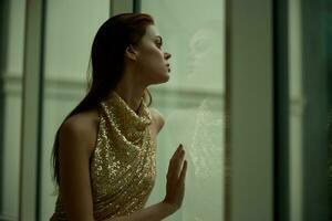 vrouw in goud lovertjes jurk staand door de venster poseren en op zoek Bij de stad in een elegant kijken met lang haar, ontwerper jurk met Open rug, partij kijken foto