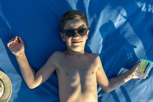 gelukkig jongen in zonnebril met kaal torso zonnen aan het liegen Aan de strand foto