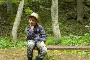 grappig Kaukasisch jongen in een reizigers hoed en windjak zit Aan een bank foto