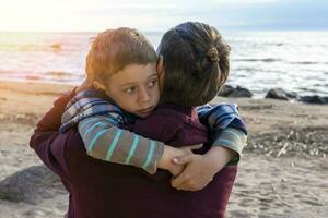 schattig verdrietig peuter- jongen knuffels zijn vader Aan de kust in zonsondergang licht foto