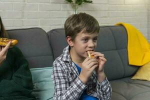 jongen is zittend Bij huis Aan de bank, aan het eten pizza en aan het kijken TV foto