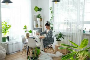 een vrouw naait tule Aan een elektrisch naaien machine in een wit modern interieur van een huis met groot ramen, huis planten. comfort in de huis, een huisvrouw hobby foto