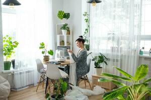een vrouw naait tule Aan een elektrisch naaien machine in een wit modern interieur van een huis met groot ramen, huis planten. comfort in de huis, een huisvrouw hobby foto