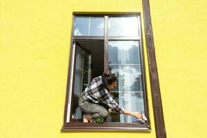 een vrouw handmatig wast de venster van de huis met een vod met een verstuiven schoonmaakster en een dweilen buiten. veiligheid Bij hoogte, herstellen bestellen en netheid in de lente, schoonmaak onderhoud foto