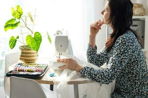 vrouw denken, dromen en naait tule Aan een elektrisch naaien machine in een wit modern interieur van een huis met groot ramen, huis planten. comfort in huis, huisvrouw hobby foto