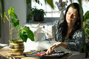 een vrouw naait tule Aan een elektrisch naaien machine in een wit modern interieur van een huis met groot ramen, huis planten. comfort in de huis, een huisvrouw hobby foto