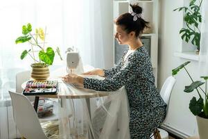 een vrouw naait tule Aan een elektrisch naaien machine in een wit modern interieur van een huis met groot ramen, huis planten. comfort in de huis, een huisvrouw hobby foto