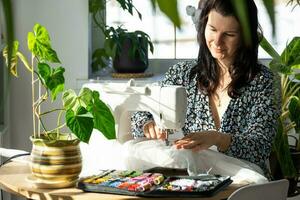 een vrouw naait tule Aan een elektrisch naaien machine in een wit modern interieur van een huis met groot ramen, huis planten. comfort in de huis, een huisvrouw hobby foto