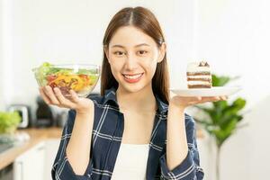 eetpatroon Aziatisch jong vrouw verliezen gewicht voor Gezondheid kiezen naar eten groente salade in schaal, verward Doen niet Kiezen eten chocola taart, bakkerij omdat zullen maken dik wanneer hongerig, vrouw gewicht verlies persoon. foto