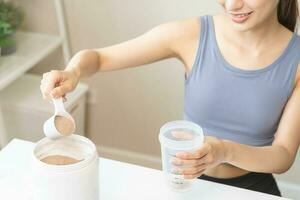 eetpatroon maaltijd vervanging voor gewicht verlies, glimlach Aziatisch jong vrouw, meisje in sportkleding, hand- in Holding lepel maken eiwit schudden, drinken supplement voor spier na training Bij huis. gezond lichaam zorg. foto