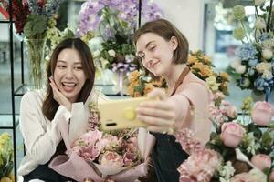 e-commerce bedrijf partner, twee jong vrouw bloemist arbeiders tonen bloemen regeling, online leven streamen, en selfies met smartphone toepassing in helder bloem winkel, mooi bloesems op te slaan. foto