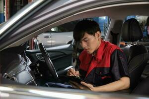 een jong deskundige Aziatisch mannetje automotive monteur technicus controle een onderhoud lijst met tablet in auto interieur Bij garage. voertuig onderhoud fix en reparatie werken, industrieel bezetting bedrijf banen. foto