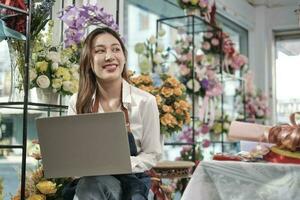 een jong Aziatisch vrouw bloemist en mkb ondernemer in schort, werken met laptop, online verkoop bloemen regelen in kleurrijk bloem winkel op te slaan met mooi bundel van bloesems, en e-commerce bedrijf. foto