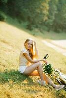 een mooi jong vrouw met een boeket van wilde bloemen is zittend Aan de gras De volgende naar haar fiets. zomer vakantie in de land foto
