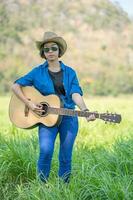 vrouw slijtage hoed en dragen haar gitaar in gras veld- foto