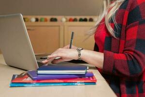 blonde vrouw achter een laptop zitten en schrijven. er ligt een laptop, een tablet, een telefoon en een notebook op tafel. foto