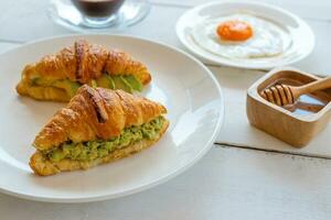 croissant belegd broodje met ei, avocado Aan wit schotel en honing, koffie. ontbijt en gezond voedsel concept. foto