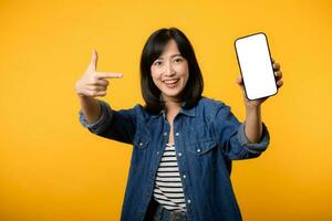 portret mooi jong Aziatisch vrouw gelukkig glimlach gekleed in denim jasje tonen smartphone scherm met richten vinger hand- gebaar naar mobiel telefoon isoleren Aan geel studio achtergrond. foto