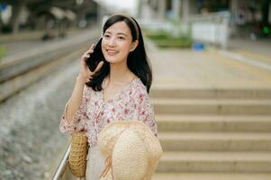 Aziatisch jong vrouw reiziger met het weven hoed gebruik makend van een mobiel telefoon naast spoorweg trein station in Bangkok. reis reis levensstijl, wereld reizen ontdekkingsreiziger of Azië zomer toerisme concept. foto
