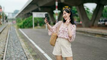 Aziatisch jong vrouw reiziger met het weven mand gebruik makend van een mobiel telefoon naast spoorweg trein station in Bangkok. reis reis levensstijl, wereld reizen ontdekkingsreiziger of Azië zomer toerisme concept. foto