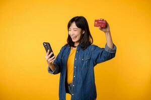 portret mooi jong Aziatisch vrouw gelukkig glimlach gekleed in denim jasje tonen smartphone en credit kaart isoleren Aan geel studio achtergrond. boodschappen doen online smartphone toepassing concept foto