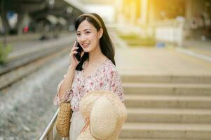Aziatisch jong vrouw reiziger met het weven hoed gebruik makend van een mobiel telefoon naast spoorweg trein station in Bangkok. reis reis levensstijl, wereld reizen ontdekkingsreiziger of Azië zomer toerisme concept. foto