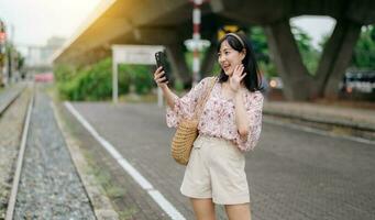 Aziatisch jong vrouw reiziger met het weven mand gebruik makend van een mobiel telefoon naast spoorweg trein station in Bangkok. reis reis levensstijl, wereld reizen ontdekkingsreiziger of Azië zomer toerisme concept. foto