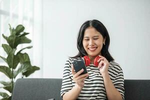portret jong Aziatisch vrouw gebruik makend van slim mobiel telefoon met koptelefoon voor luister muziek- Aan sofa foto