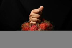Aziatisch mannetje donker huid single hand- vuist vinger Aan zwart achtergrond Holding ramboetan fruit foto