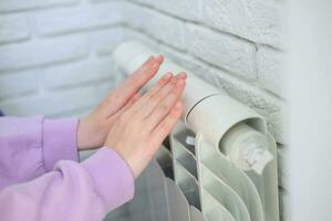 de kind handen warm hun handen in de buurt de verwarming radiator. besparing gas- in de verwarming seizoen. foto