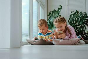 kinderen liggen Aan de verdieping van de venster en kijk maar tekenfilms Aan hun telefoons. broer en zus Speel spellen Aan de telefoon. foto