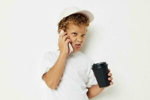 schattig weinig jongen in een wit t-shirt pet met een telefoon in een glas met een drinken licht achtergrond ongewijzigd foto