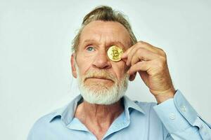 een ouderen Mens in een blauw overhemd covers de ogen van een bitcoin munt foto