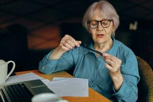 portret van een ouderen vrouw met bril zit Bij een tafel in voorkant van een laptop freelancer werken ongewijzigd foto