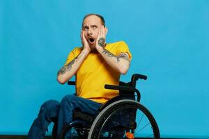 een Mens in een rolstoel verrast en Open mond in een t-shirt met tatoeages Aan zijn armen zit Aan een blauw studio achtergrond, een vol leven, een echt persoon foto