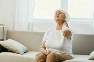 ouderen vrouw erge, ernstige nek pijn zittend Aan de bank, Gezondheid problemen in oud leeftijd, arm kwaliteit van leven. foto