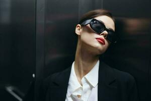 elegant vrouw in een zwart jasje en zonnebril poseren in een lift optillen in de buurt de spiegel, mode model, donker filmische licht en kleur Matrix, aantrekkingskracht wijnoogst foto