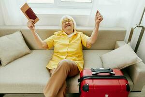 gelukkig senior vrouw met paspoort en reizen ticket Ingepakt een rood koffer, vakantie en Gezondheid zorg. glimlachen oud vrouw blij zittend Aan de sofa voordat de reis verheven haar handen omhoog in vreugde. foto