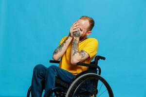 een Mens in een rolstoel glimlach looks Bij de camera in een t-shirt met tatoeages Aan zijn armen zit Aan een blauw studio achtergrond, een vol leven, een echt persoon foto