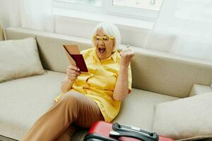 gelukkig senior vrouw met paspoort en reizen ticket Ingepakt een rood koffer, vakantie en Gezondheid zorg. glimlachen oud vrouw blij zittend Aan de sofa voordat de reis verheven haar handen omhoog in vreugde. foto